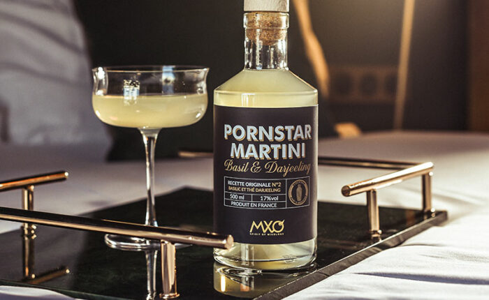 Bouteille du Cocktail Pornstar Martini MXO Spirit of Mixology posée sur un plateau avec un verre sur un lit