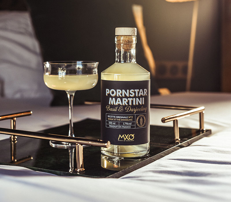 Bouteille du Cocktail Pornstar Martini MXO Spirit of Mixology posée sur un plateau avec un verre sur un lit